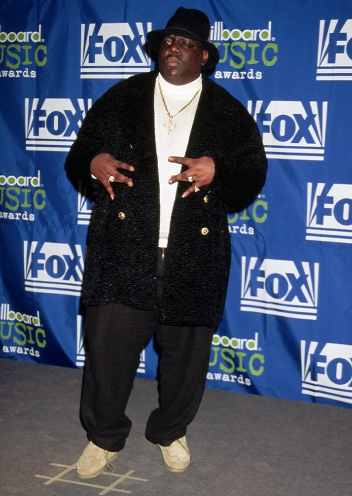 Homem vestindo casaco preto e chapéu na premiação, usando tênis estilo Wallabees. Fundo com logotipos da Fox e Billboard Music Awards.