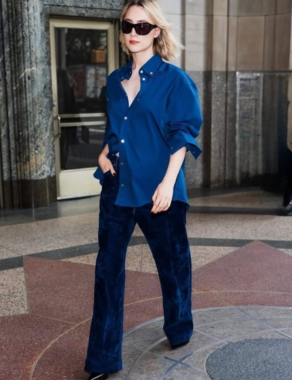 Saoirse Ronan está com uma camisa azul e óculos escuros, andando em frente a um edifício.