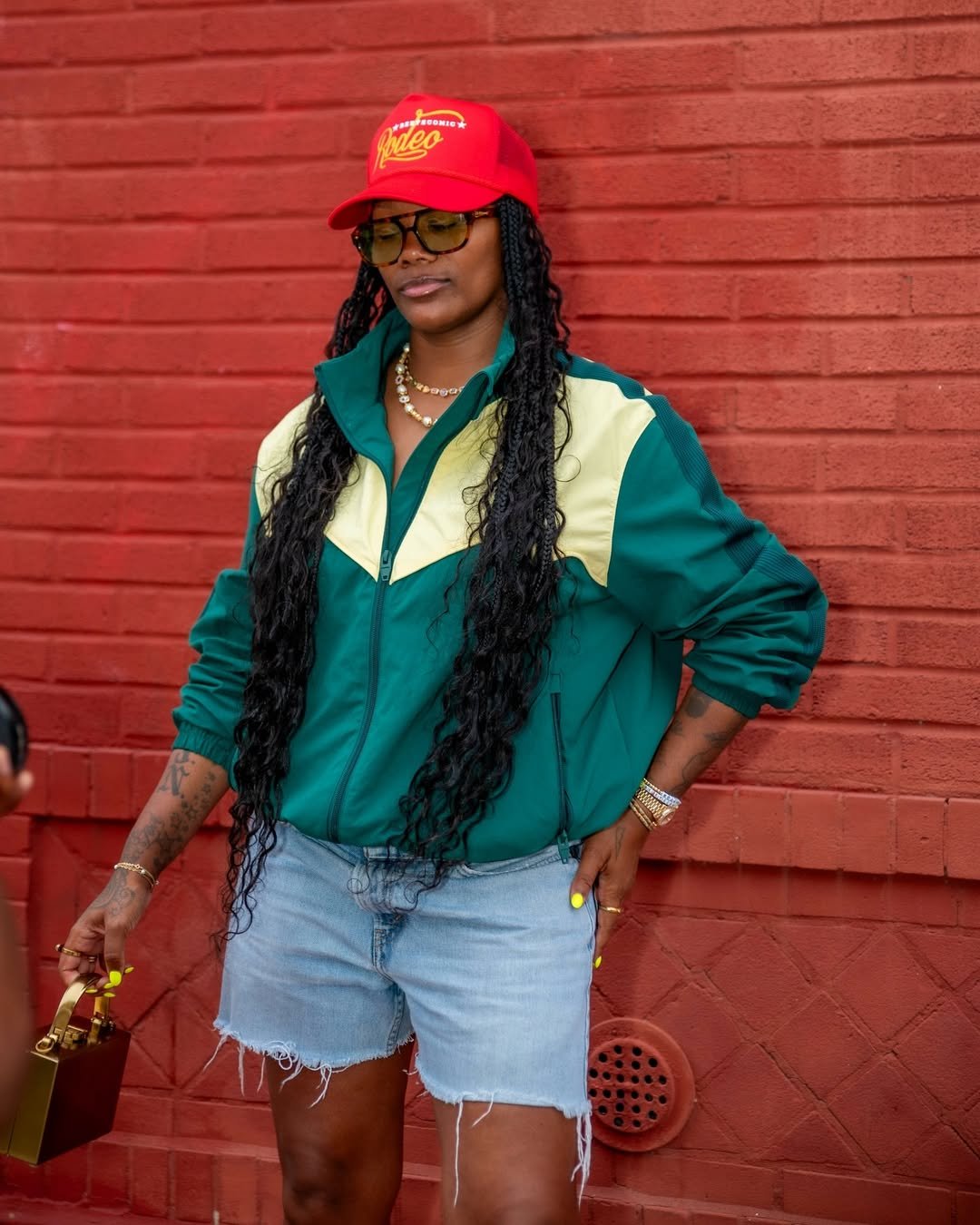 Mulher de boné vermelho, óculos escuros e relógio dourado. Veste jaqueta verde e shorts jeans, encostada em parede vermelha.