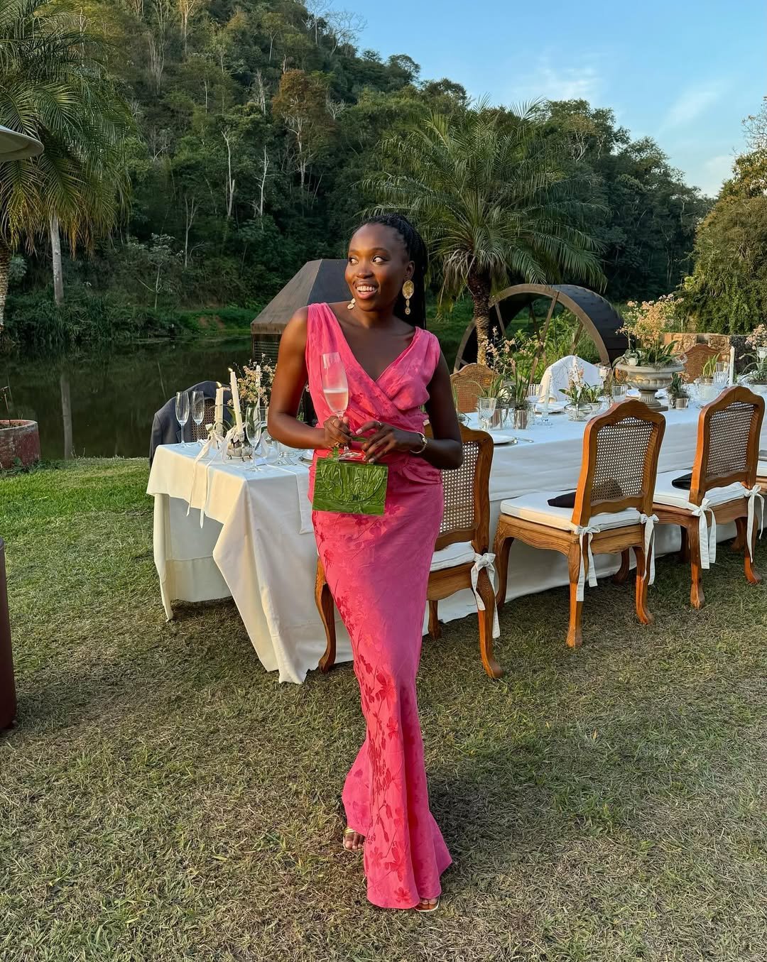 Mulher em vestido rosa segura taça de champanhe ao ar livre, mesa elegante ao fundo. Ela usa um relógio dourado discreto.