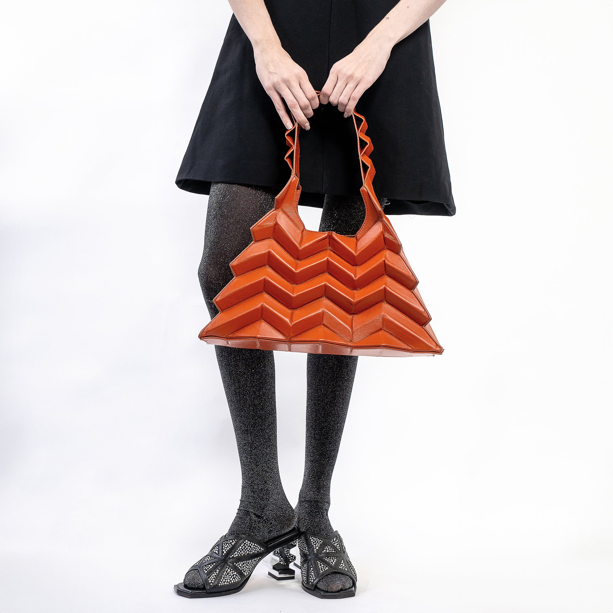 Bolsa laranja em formato geométrico, combinada com vestido preto e sandálias decoradas, destacando collabs de moda e beleza.