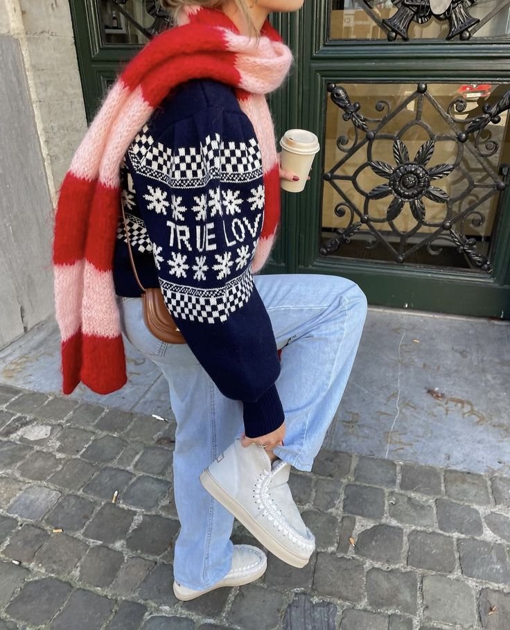 Pessoa com suéter azul de flocos de neve e frase, calças jeans e cachecol vermelho, segurando café.