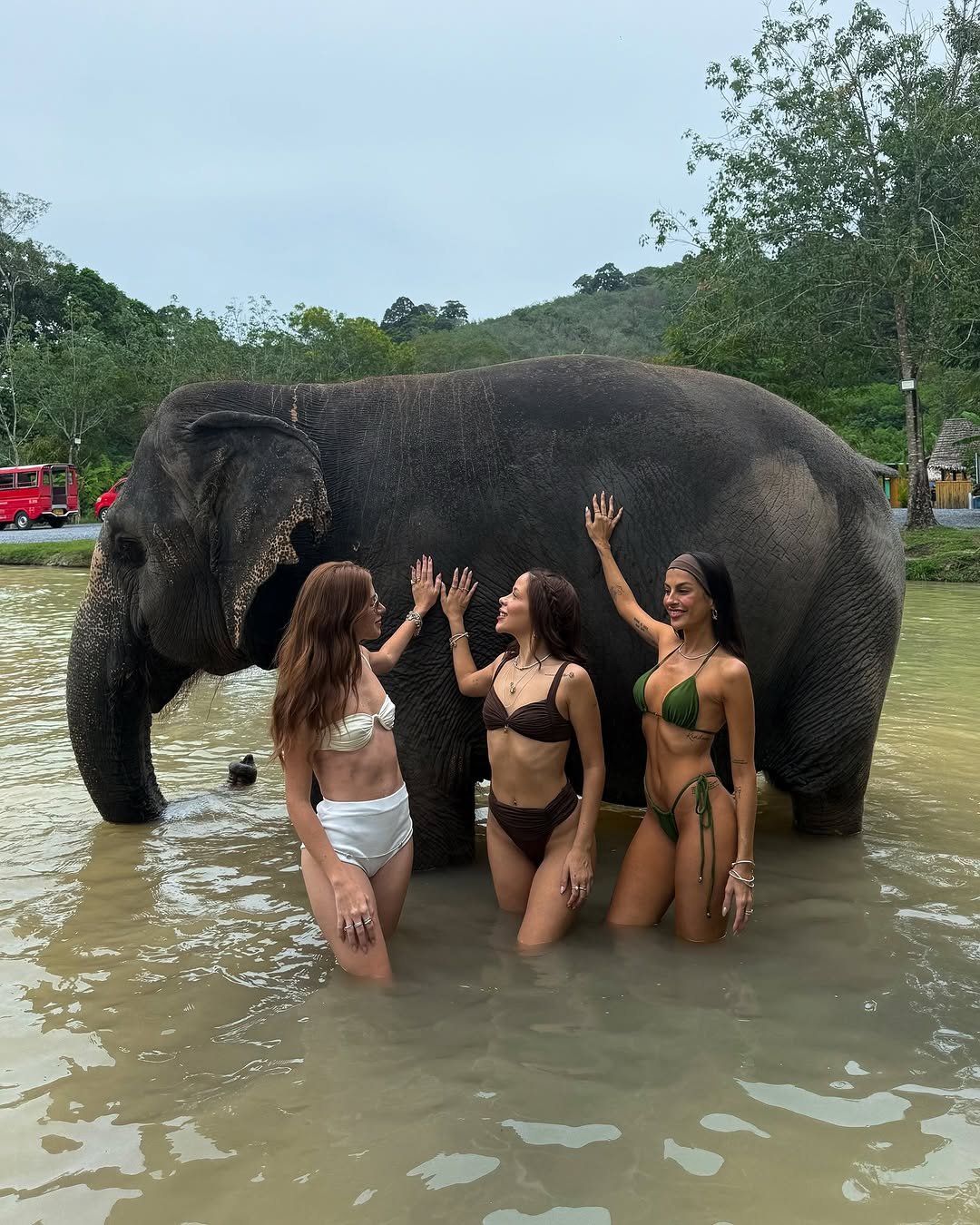 Mulheres em trajes de banho interagem com um elefante em um rio, em cenário tropical na Tailândia.