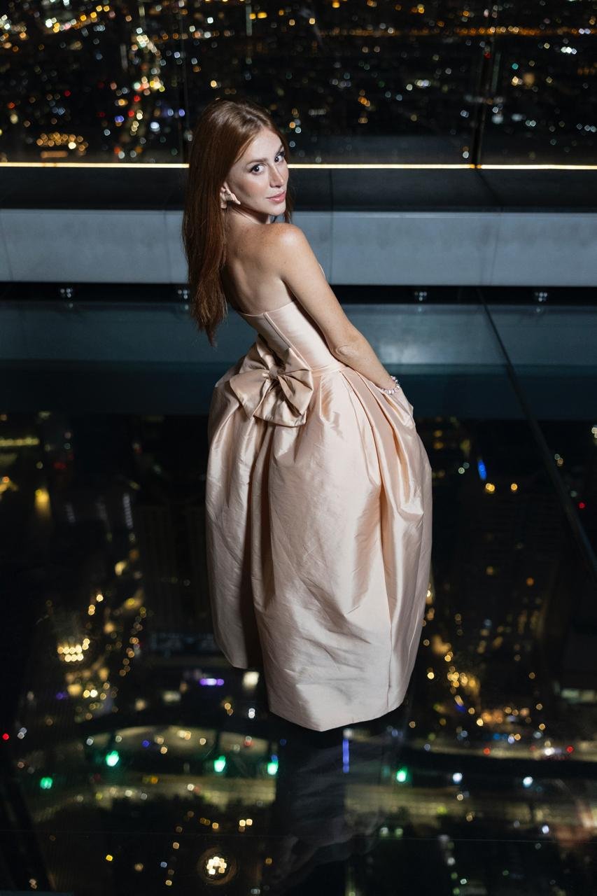 Pessoa em vestido elegante em varanda com vista panorâmica de cidade iluminada à noite. Ambiente lembra grandes cidades como as da Tailândia.