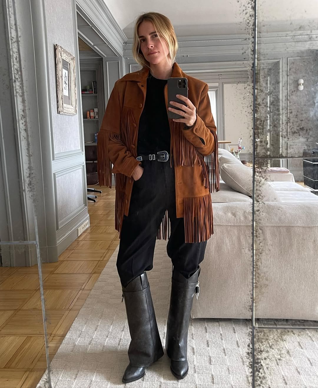 Casaco com franjas estilo anos 70, calça escura e botas. Mulher tirando selfie em sala elegante.