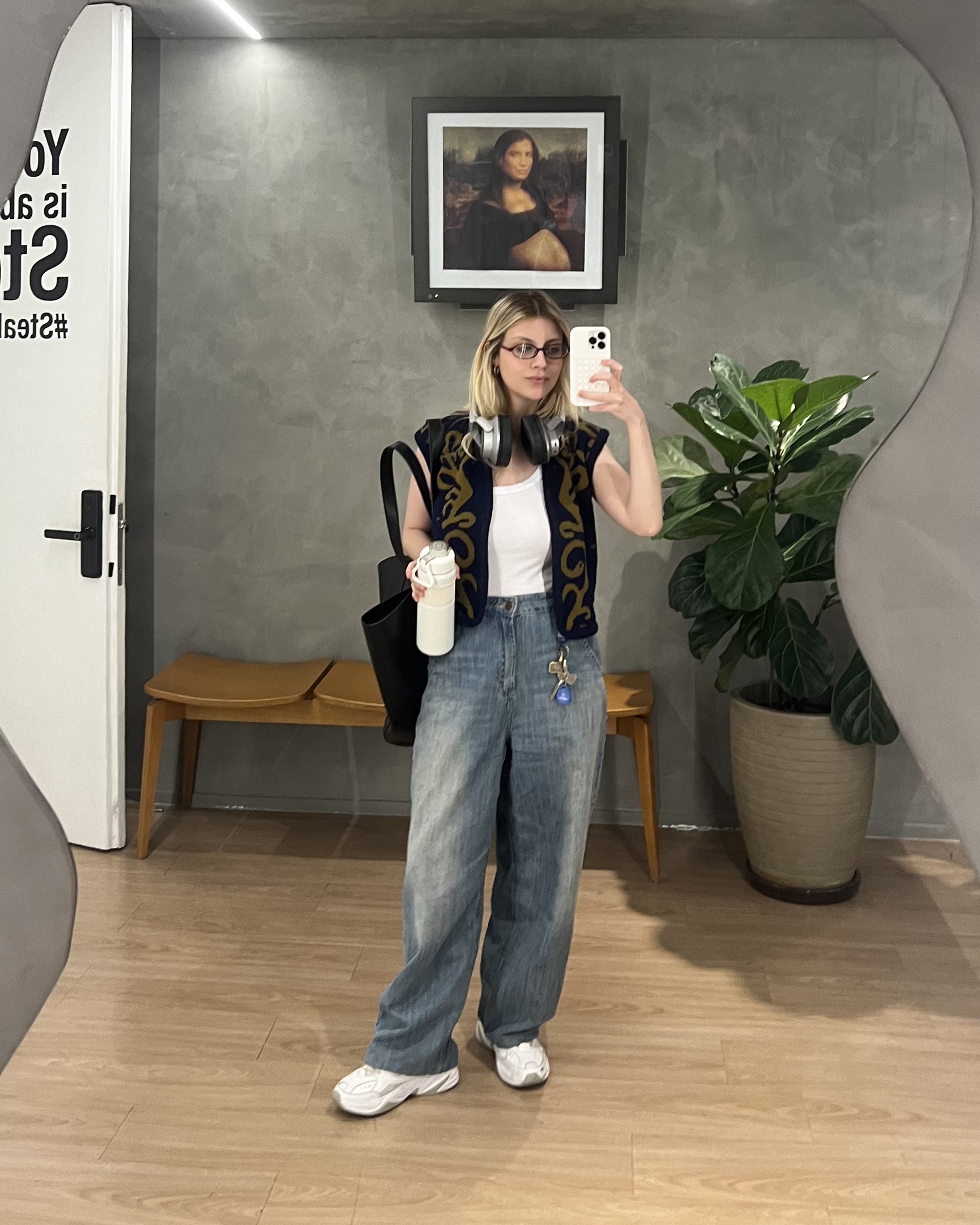 Mulher com calça jeans e colete posando em espelho. Planta e quadro ao fundo. 