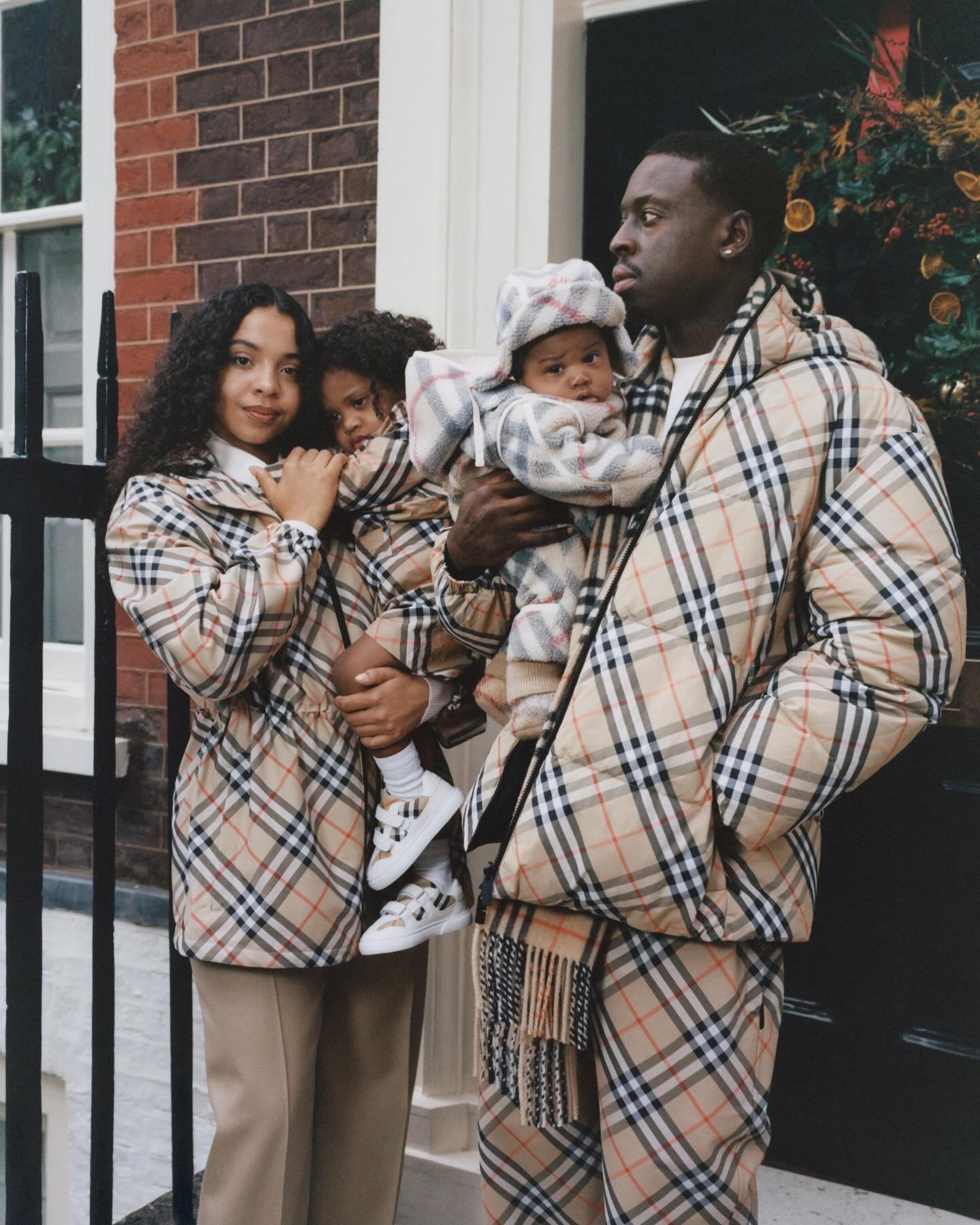 Família vestindo roupas com padrão quadriculado em frente a uma casa com decoração natalina.