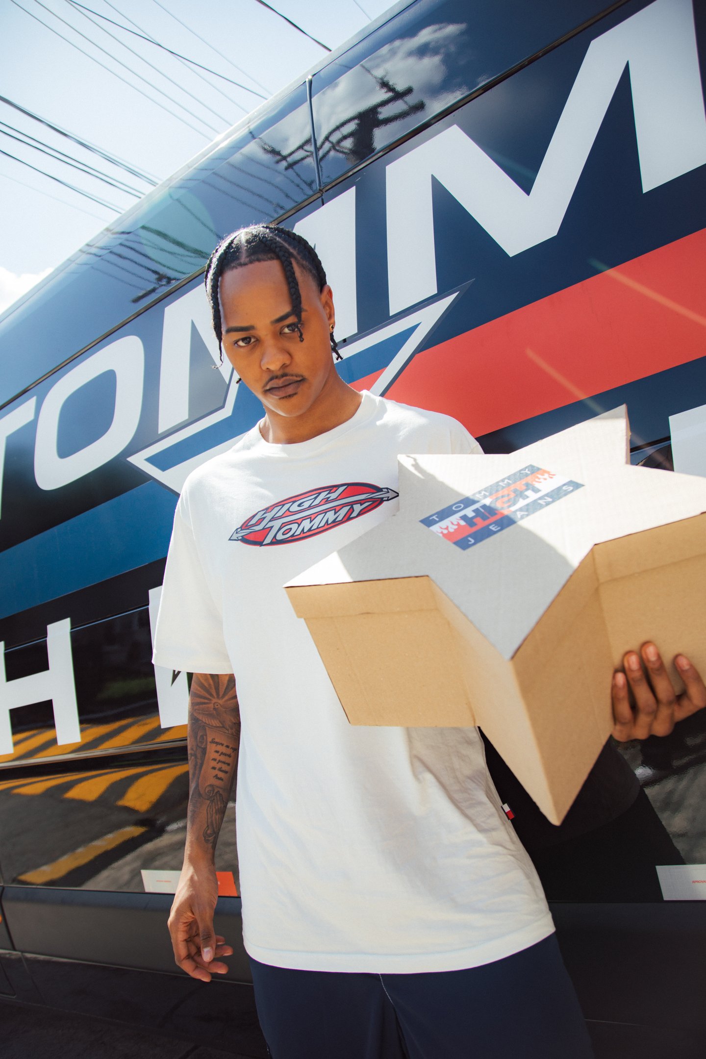 Homem com camiseta branca segura uma caixa na frente de uma van estampada; influência de collabs de moda e beleza.