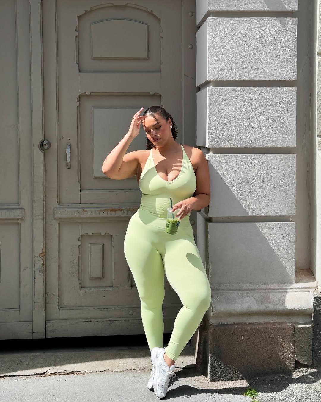 Mulher de roupa fitness verde clara em frente a uma porta, segurando bebida. O alt text deve incluir tops de academias.