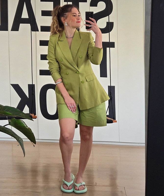 Mulher com blazer verde e shorts, usando chinelo estiloso verde, tira selfie em frente a espelho.