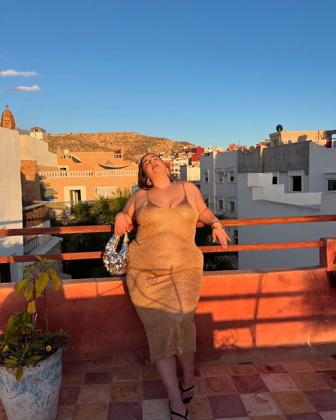 Mulher usando vestido de alcinha dourado em terraço ensolarado, segurando uma bolsa brilhante.