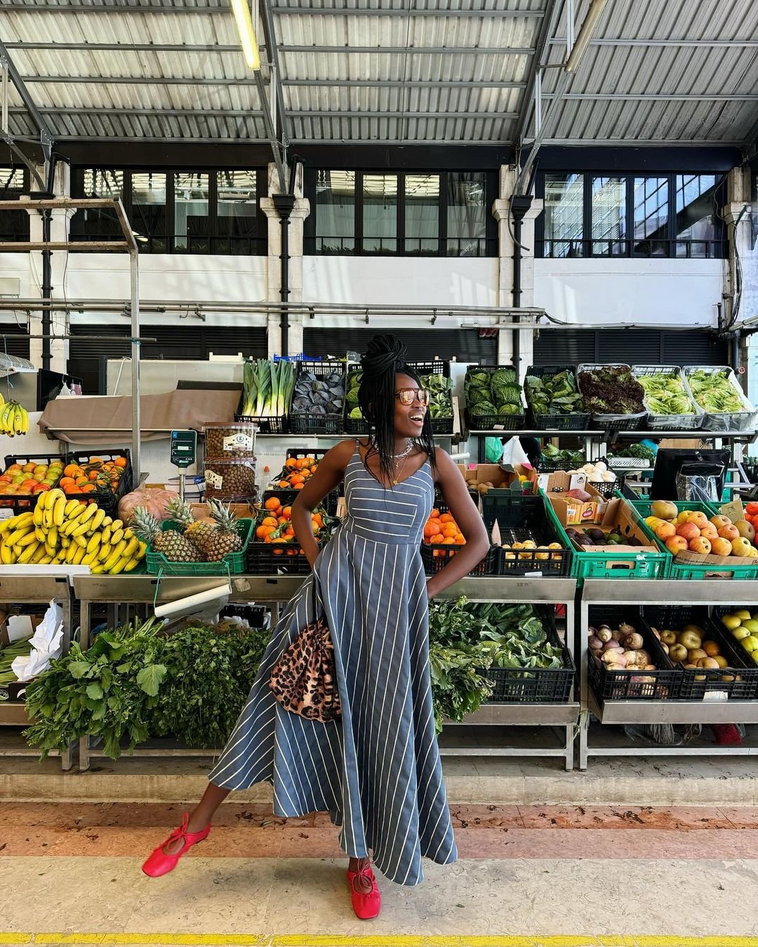 Mulher posando em um mercado de frutas e verduras, usando um vestido de alcinha listrado e sapatos vermelhos.