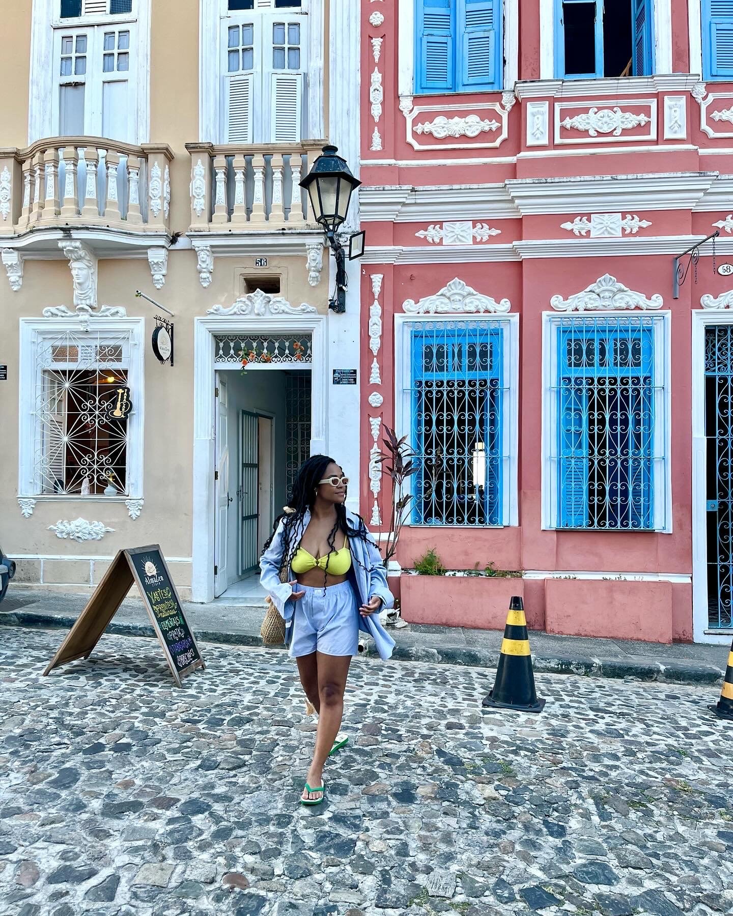 Pessoa caminha por rua de pedras em Salvador, com fachadas coloridas ao fundo. Ela usa roupas leves e carrega uma bolsa.