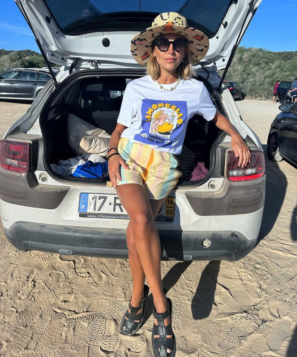 Pessoa com chapéu de sol, óculos escuros e roupas casuais sentada na traseira de um carro na praia.