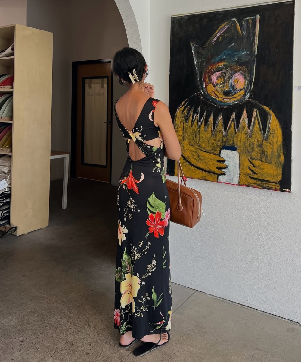 Pessoa em um vestido floral observando uma pintura em uma galeria, com uma bolsa elegante e acessórios de verão.