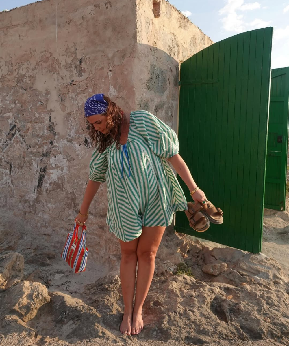 Mulher com vestido listrado e lenço, segurando bolsa e sandálias, em cenário rústico. Acessórios de verão complementam o visual.