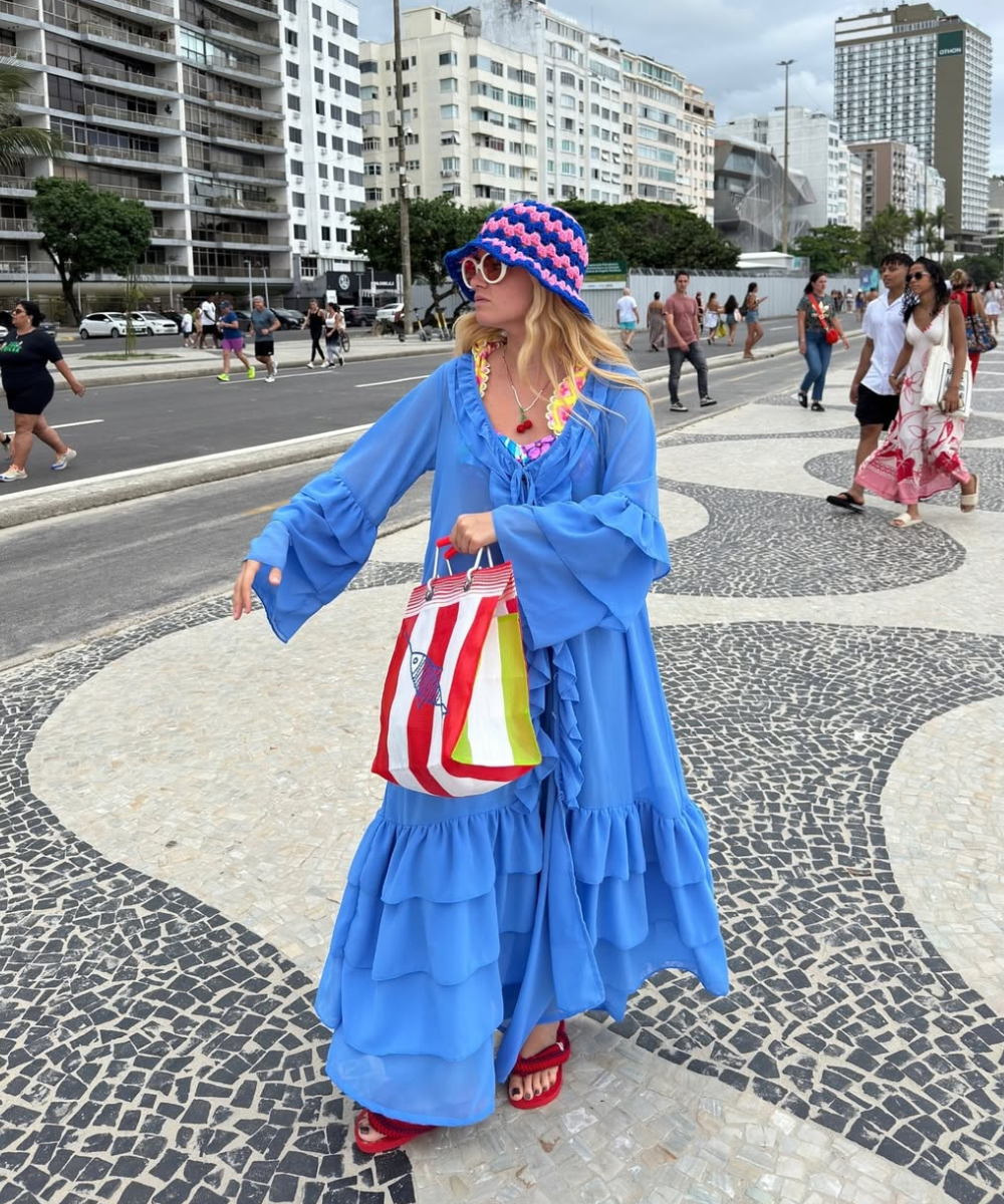 Pessoa na rua com chapéu colorido, óculos e bolsa listrada, exemplificando acessórios de verão. Fundo com prédios e pessoas.