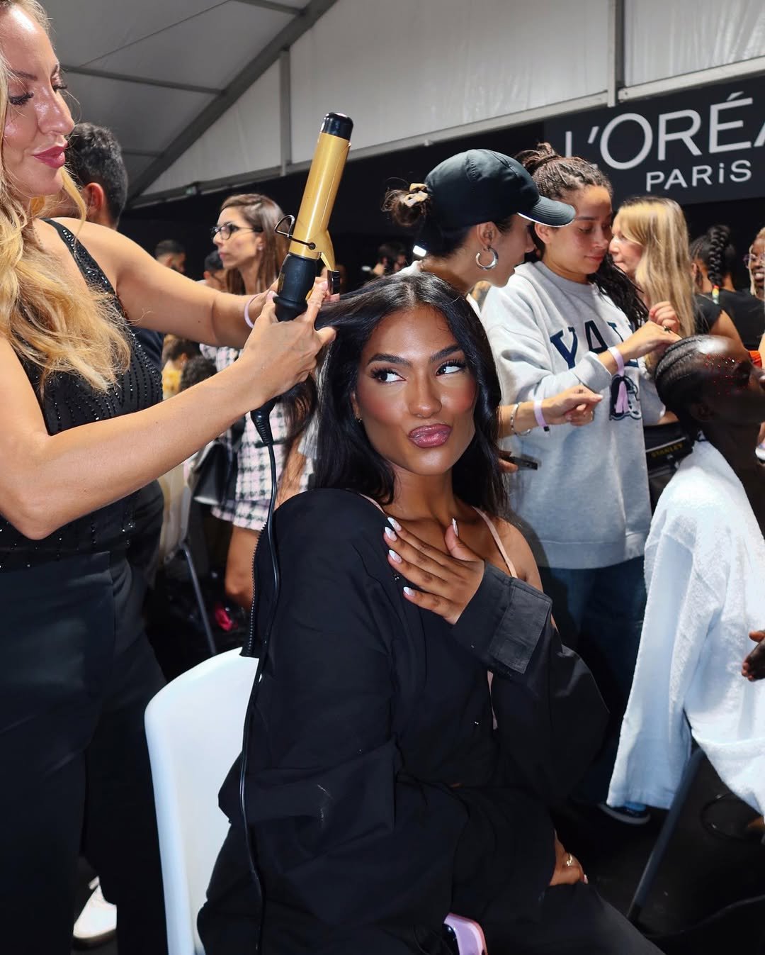 Mulher com cabelo volumoso em salão de beleza, usando chapinha; estilistas e clientes ao fundo. adeus ao cabelo fino