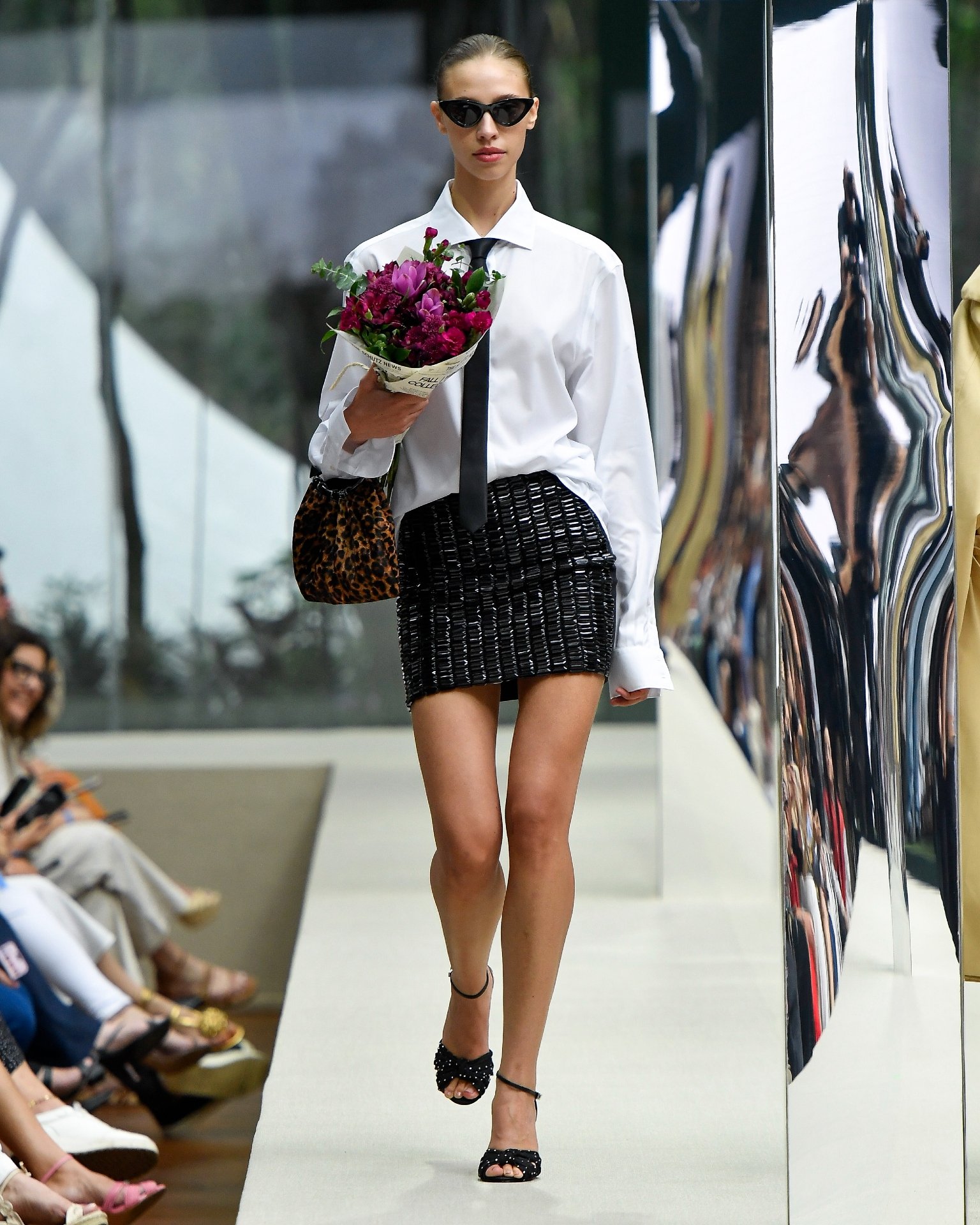 Modelo caminha na passarela em desfile da Arezzo&Co., usando camisa branca, saia preta e óculos escuros, segurando flores.