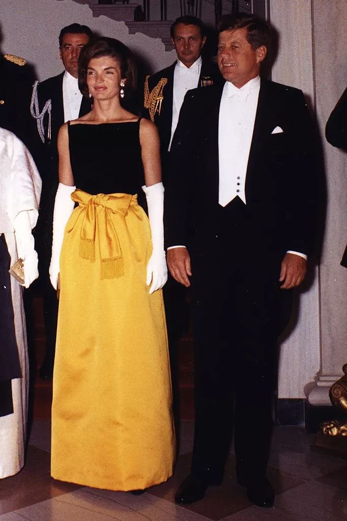 Pessoas vestidas formalmente em um evento social. Mulher com vestido amarelo e homem com smoking.