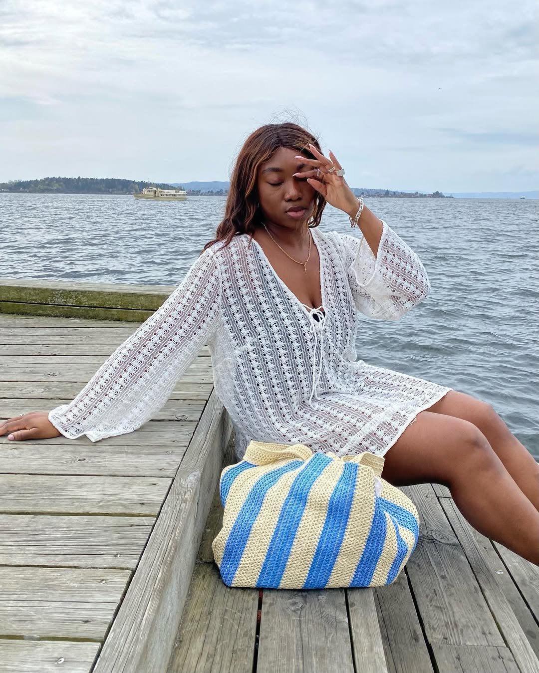 Mulher em um píer com bolsa de praia listrada ao lado, usando túnica branca na frente do mar.