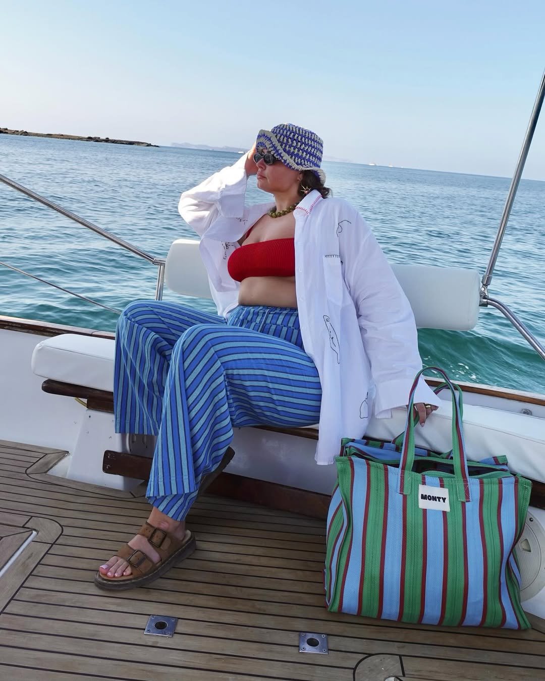 Mulher em barco com calça listrada e chapéu. Ao lado, bolsa de praia listrada verde e azul.