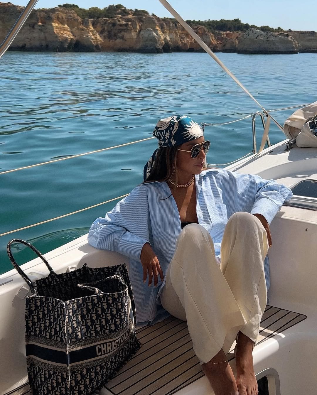 Pessoa relaxando em um barco com uma grande bolsa de praia estampada ao lado, mar azul e falésias ao fundo.