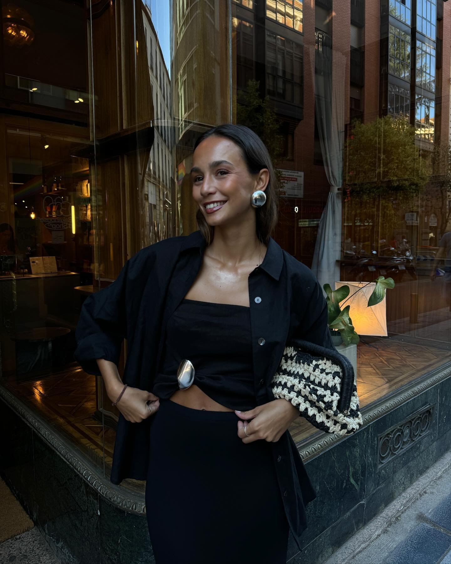 Pessoa sorrindo em frente a uma vitrine, segurando uma bolsa de ráfia preta e bege.