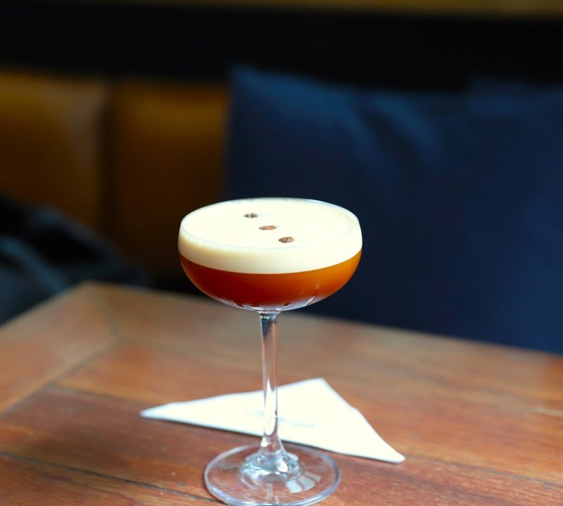 Bebida escura em taça sobre mesa de madeira, com espuma branca e grãos de café decorativos. Café gelado.