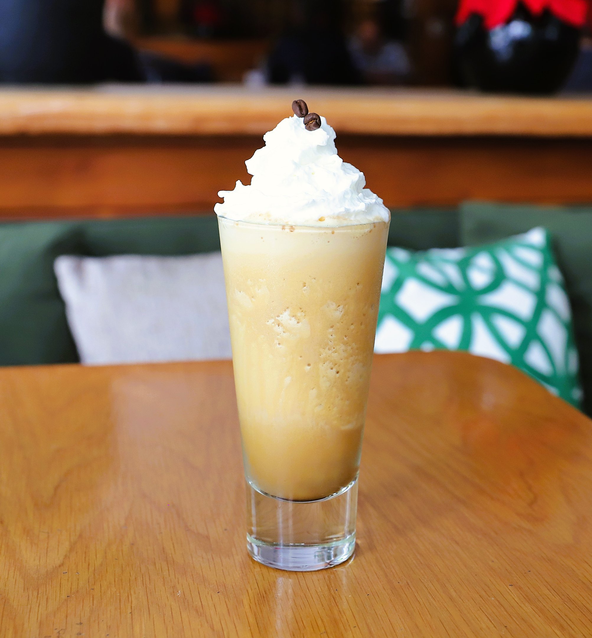 Bebida de café gelado com chantilly e grãos de café em copo alto, sobre mesa de madeira.