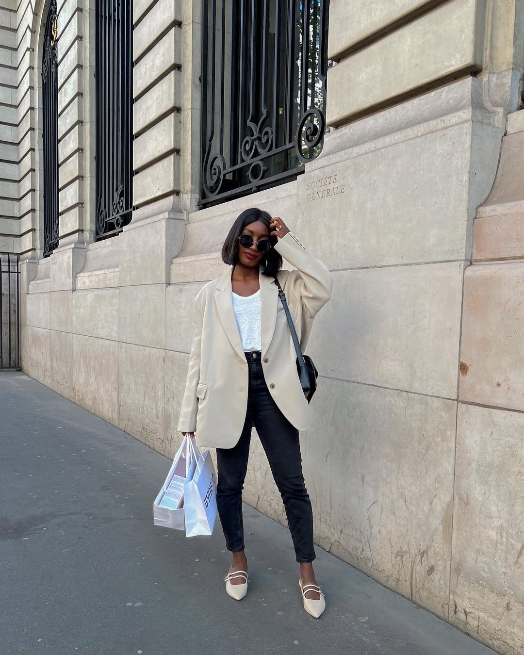 Pessoa de óculos escuros, blazer claro e calça skinny preta, segurando sacolas em frente a um edifício.
