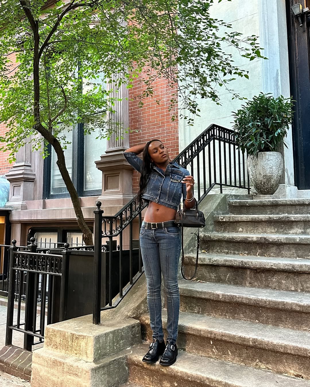 Pessoa em calça skinny jeans e jaqueta, de pé em uma escadaria ao ar livre, cercada por vegetação e edifícios de tijolos.