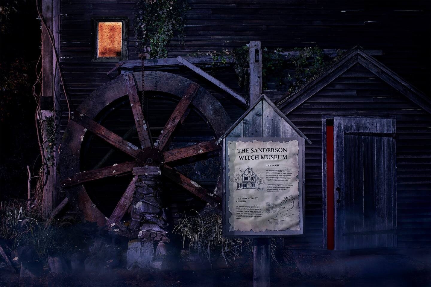 Museu de bruxas com roda de moinho, remetendo a casas temáticas. Janela iluminada e placa informativa destacam-se.