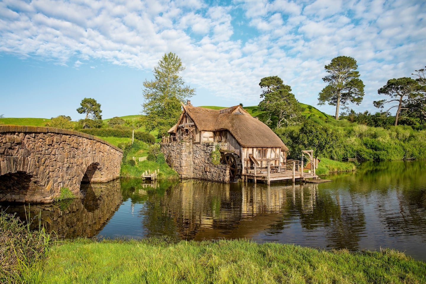 Casa temática à beira de um lago, com telhado de palha e roda d'água, ao lado de uma ponte de pedra.