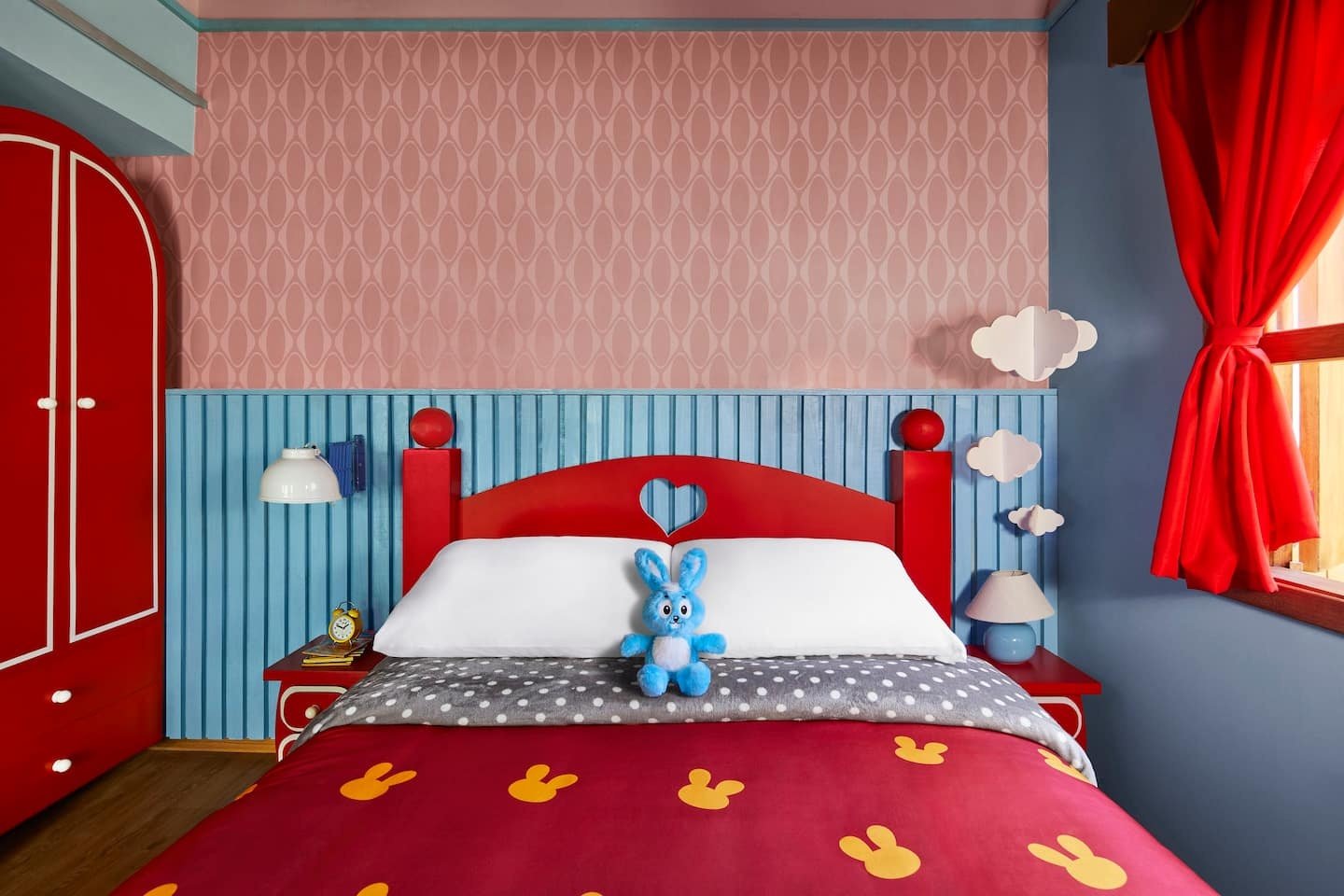 Quarto infantil colorido com cama vermelha, detalhes de nuvens e coelho de pelúcia azul. As cores vivas são características de casas temáticas.