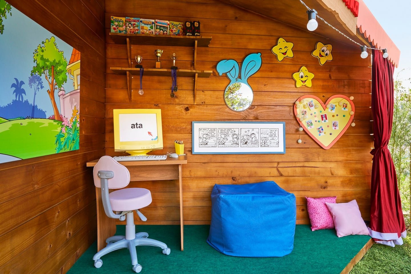 Espaço infantil em uma casa temática com mesa, computador clássico, pufes e quadrinhos na parede de madeira.