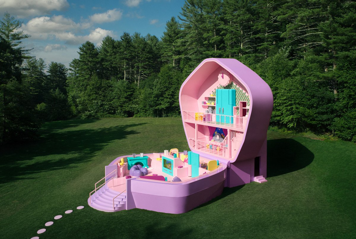 Casa temática em formato de brinquedo gigante, pintada de rosa e lavanda, em meio a uma área verde.