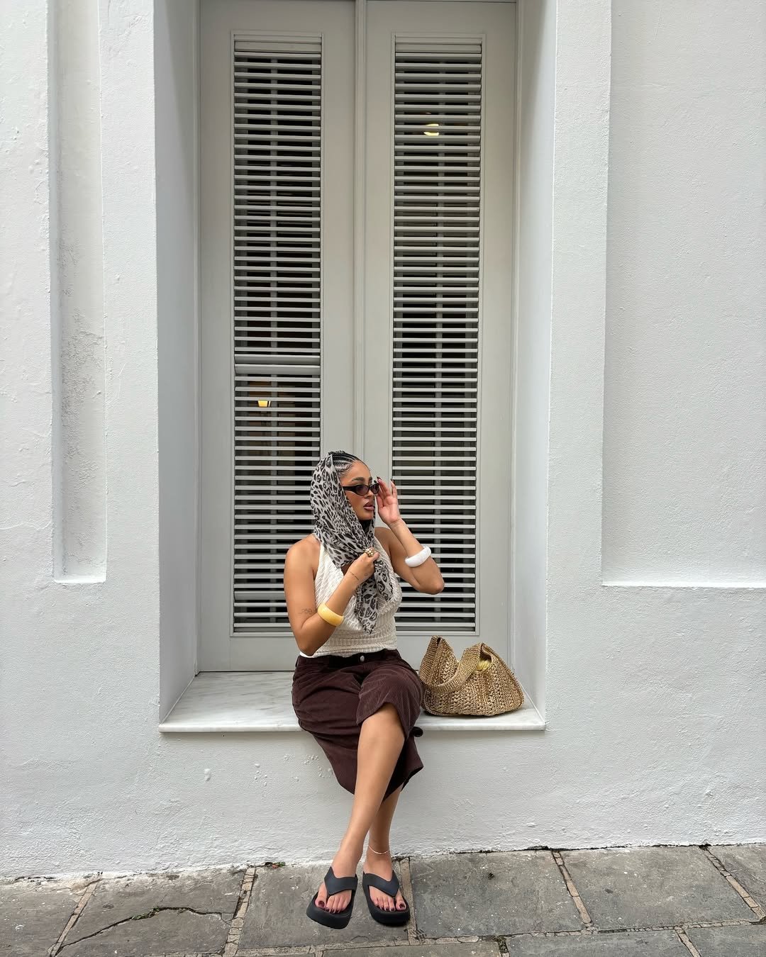 Pessoa sentada usando saia marrom e chinelo feminino preto, com bolsa e lenço de oncinha na cabeça. Fundo: parede branca e janela.