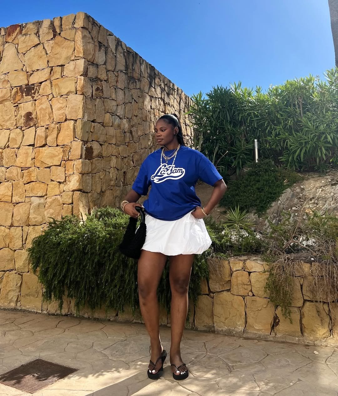 Pessoa em pé com camiseta azul, saia branca e chinelo feminino preto, em frente a parede de pedra e plantas.