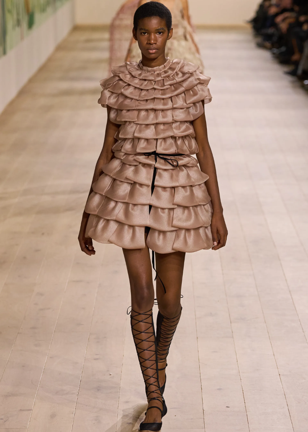 Modelo em vestido com camadas volumosas bege desfila na Semana de Alta-Costura 2025, calçando sandálias amarradas até os joelhos.