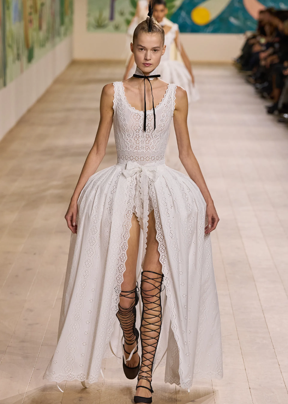 Modelo desfila com vestido branco rendado e amarração na cintura, completado por sandálias gladiadoras pretas.