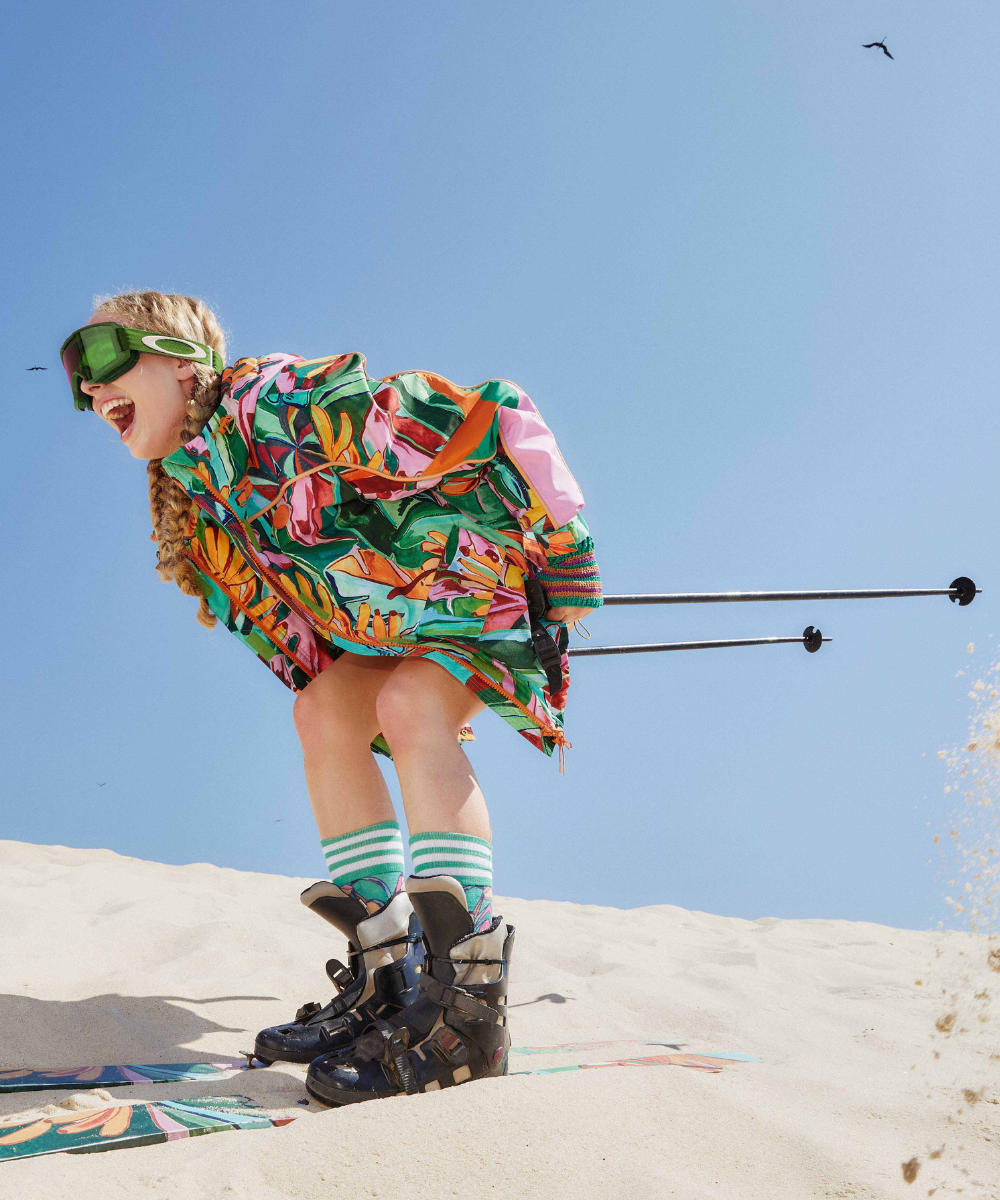 Pessoa com roupa colorida de ski e óculos de proteção desliza na areia, destacando a nova coleção de ski.