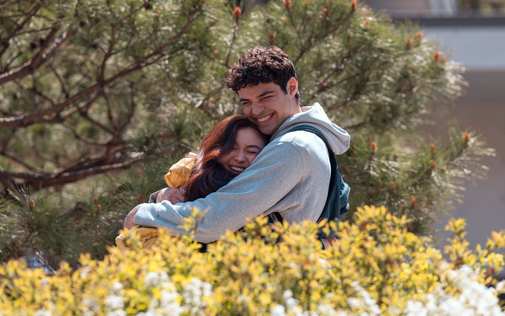 Dois jovens felizes se abraçam com carinho ao ar livre, rodeados por árvores e flores amarelas. Imagens da segunda temporada de Com Carinho, Kitty.