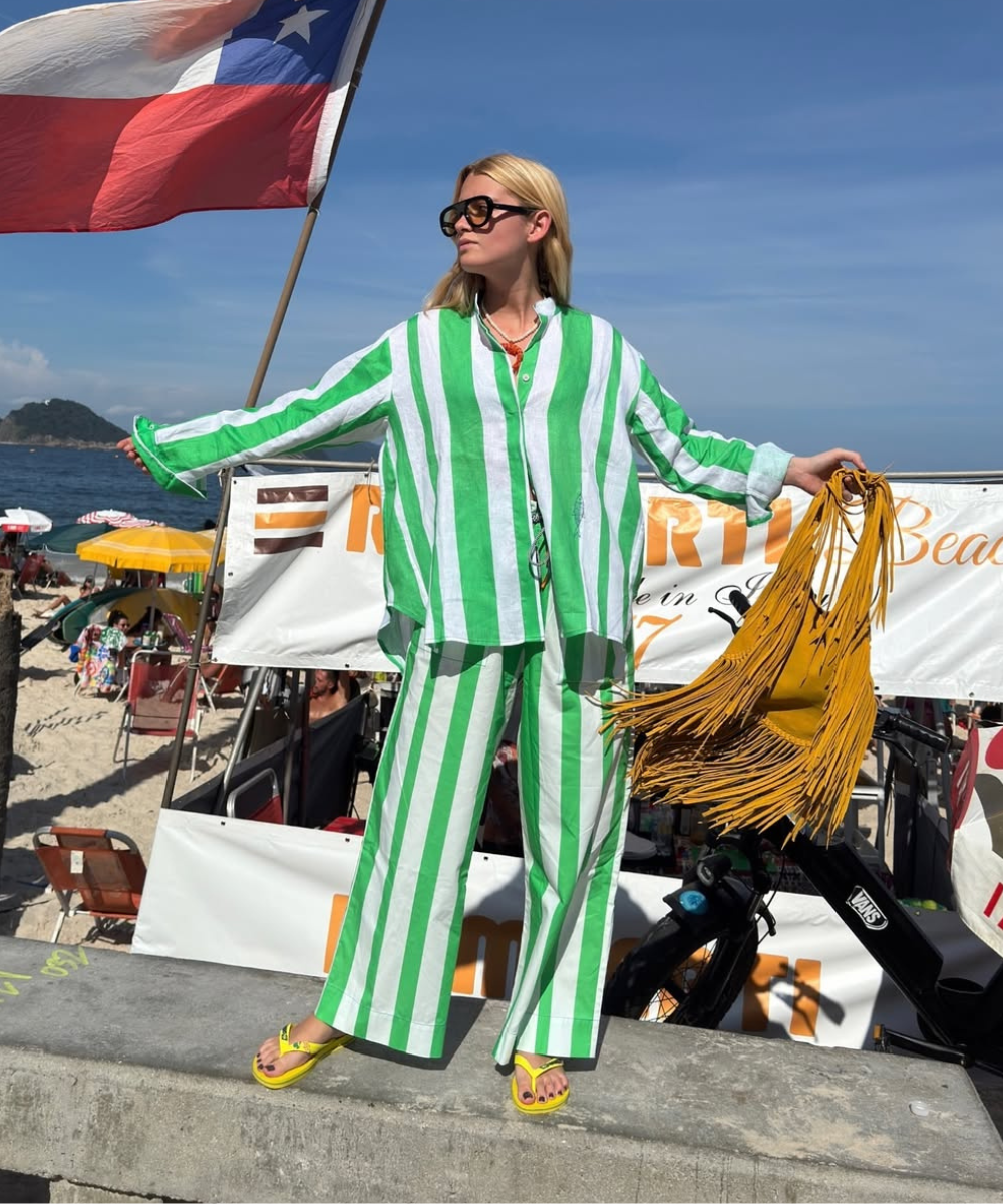 Pessoa em conjuntos estilosos listrados verde e branco, em uma praia, segurando uma bolsa amarela com franjas.