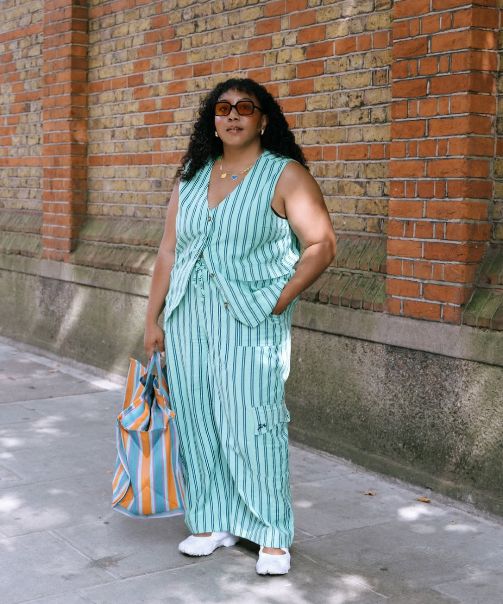 Mulher usando conjunto listrado azul e branco com bolsa combinando, posando em frente a parede de tijolos. Conjuntos estilosos.