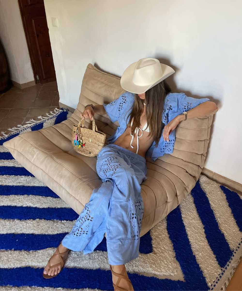 Pessoa usando chapéu branco e conjunto azul rendado, segurando bolsa de palha. Estilo casual em ambiente interno.