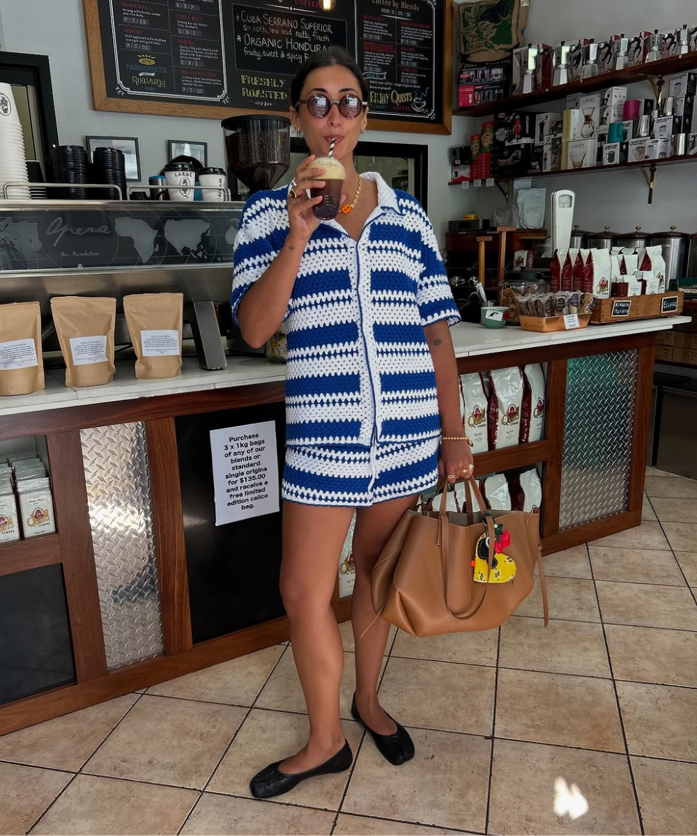 Pessoa com vestido de crochê azul e branco, segurando bolsa marrom em café. Conjuntos estilosos.