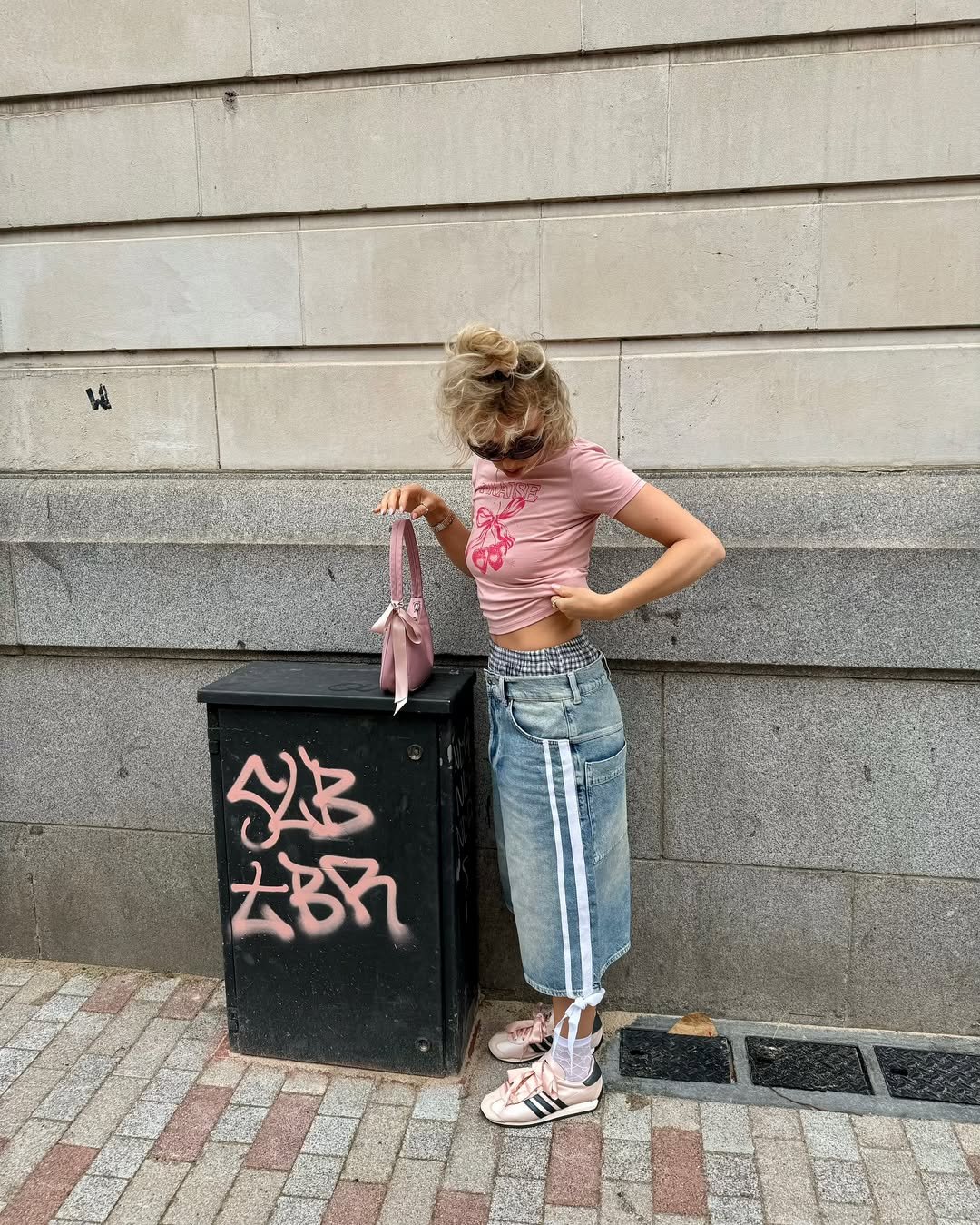 Mulher em roupa de cores pastel posa com bolsa rosa clara ao lado de uma caixa de energia grafitada na rua.