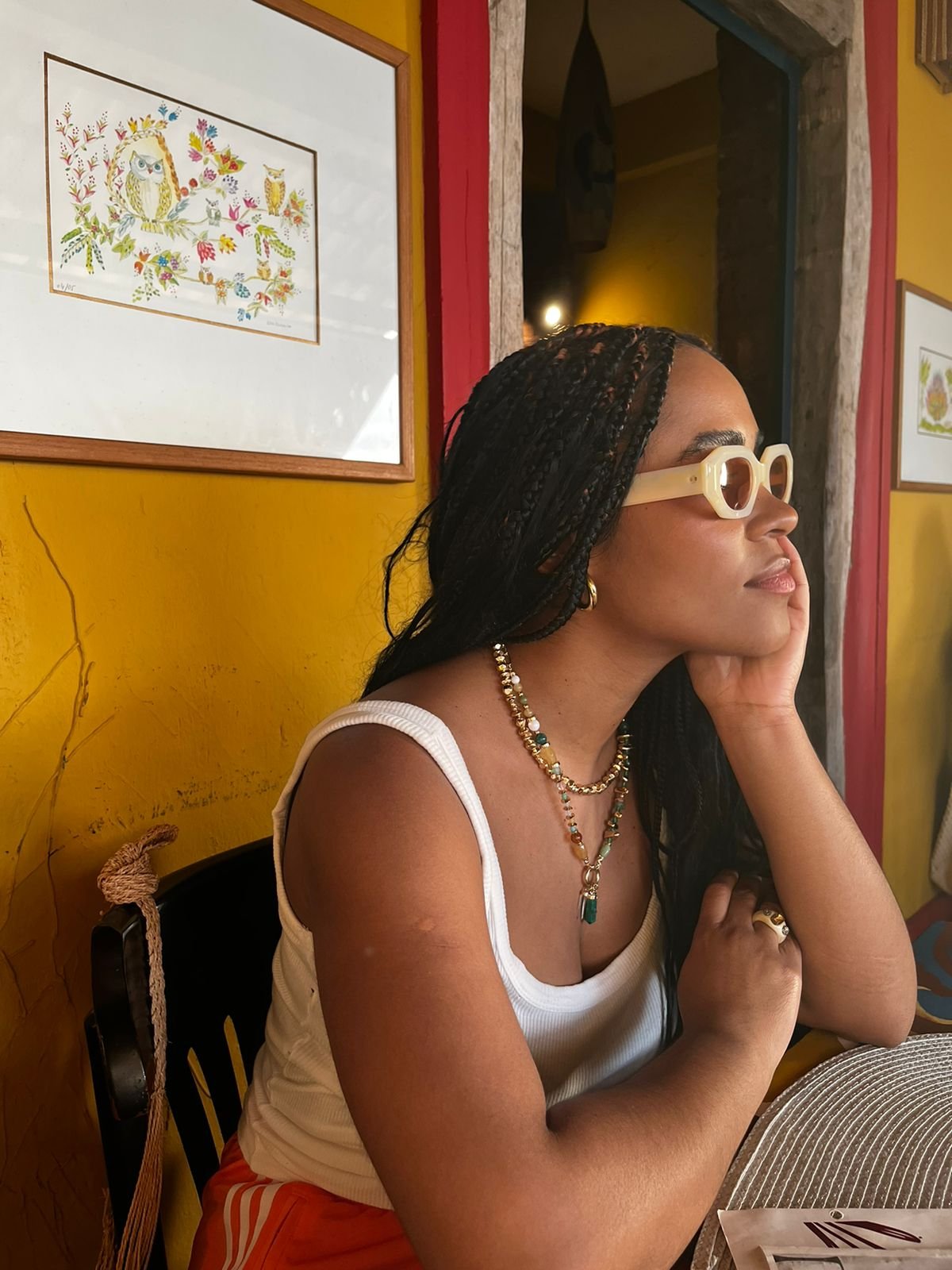 Mulher relaxa à mesa de um café colorido, exibindo um estilo dolce far niente. Ela usa óculos de sol e colar.