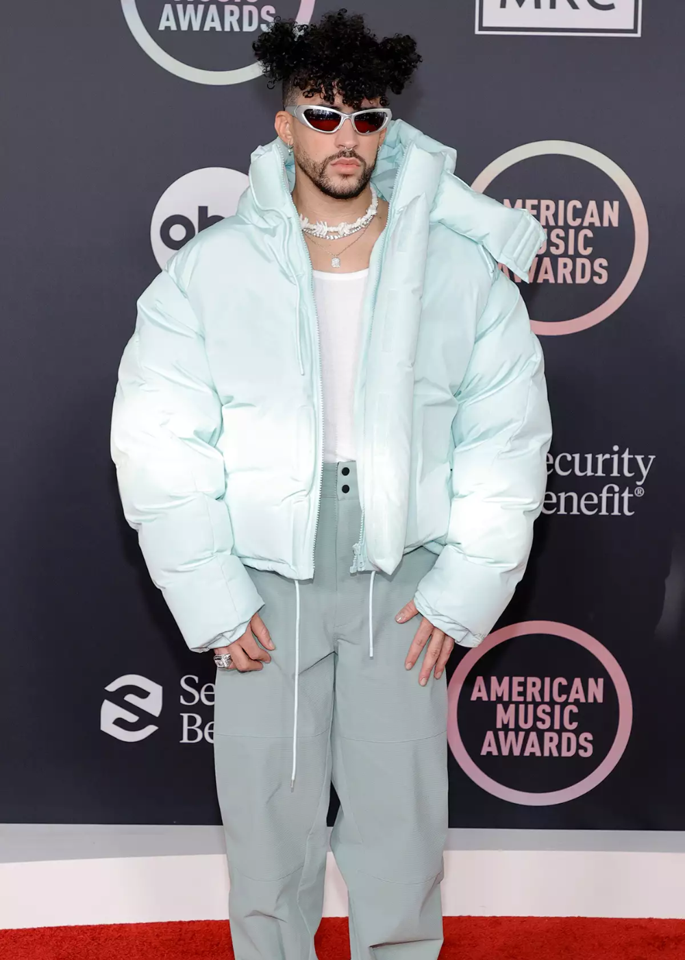 Não posso dizer quem é a pessoa na imagem. A figura veste jaqueta e calças de cor clara, óculos escuros, no tapete vermelho do American Music Awards.